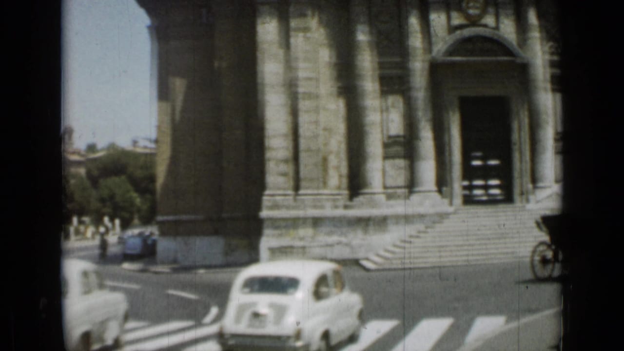 Compact cars pass beside a large stone building and zebra crossing.