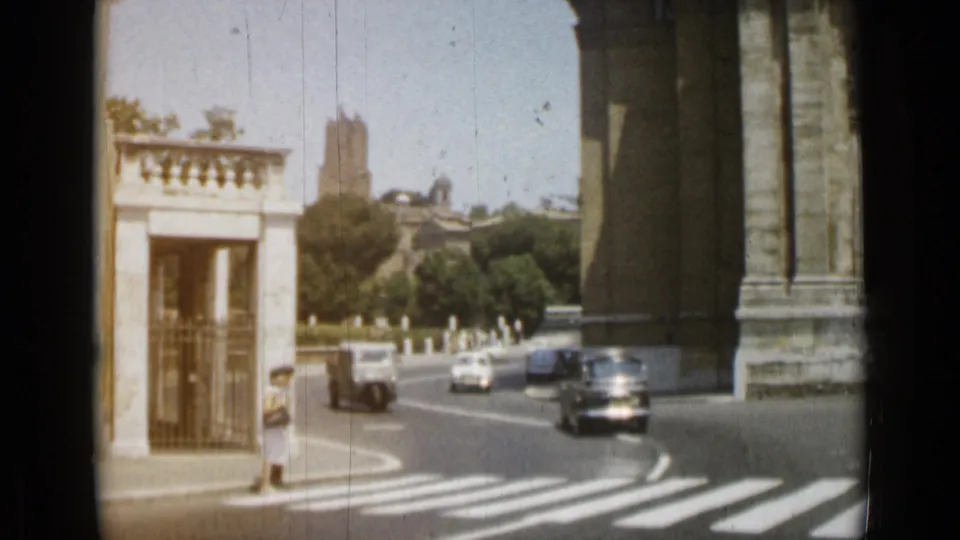 1962 Rome street view with a small booth, period cars, zebra crossing, heavy stone building edge, trees, pedestrians, and distant architecture.