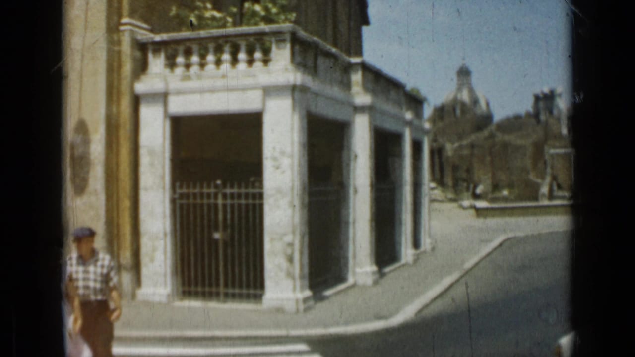 1962 Rome 8mm frame showing a white stone gate or entrance, iron railings, street curb, and a passerby in a cap.