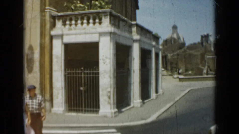 1962 Rome 8mm frame showing a white stone gate or entrance, iron railings, street curb, and a passerby in a cap.