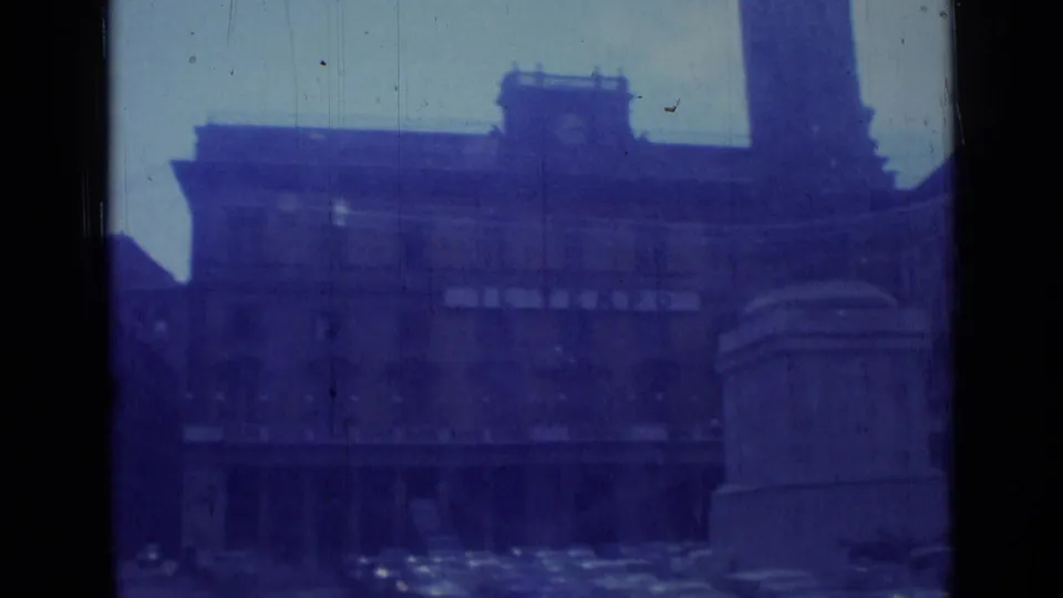 1962 Rome home-movie frame showing a clock-tower building, street traffic, and a partial monument or column from a moving car.