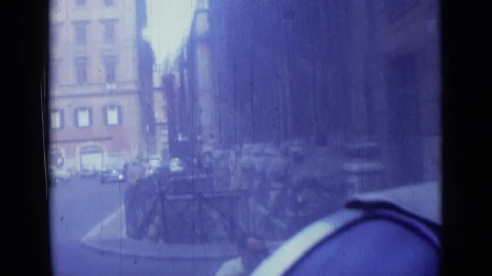 1962 Rome street view from a moving car, showing buildings, railings, traffic, and a rounded foreground car edge.