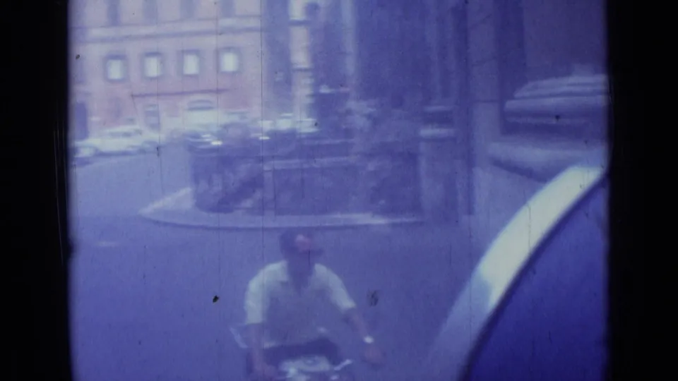 In Rome, a man on a bicycle passes through a street scene with parked cars and older building facades, seen through blue-shifted 8mm film.