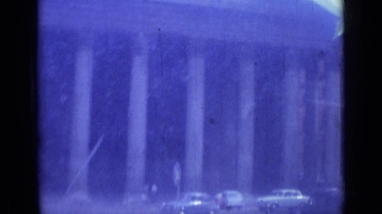 1962 Rome street view from a car, showing a grand columned facade, classic cars, and pedestrians through blue-shifted 8mm film.