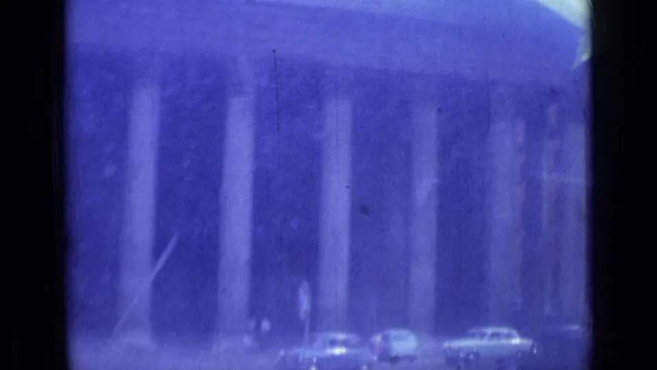 1962 Rome street view from a car, showing a grand columned facade, classic cars, and pedestrians through blue-shifted 8mm film.