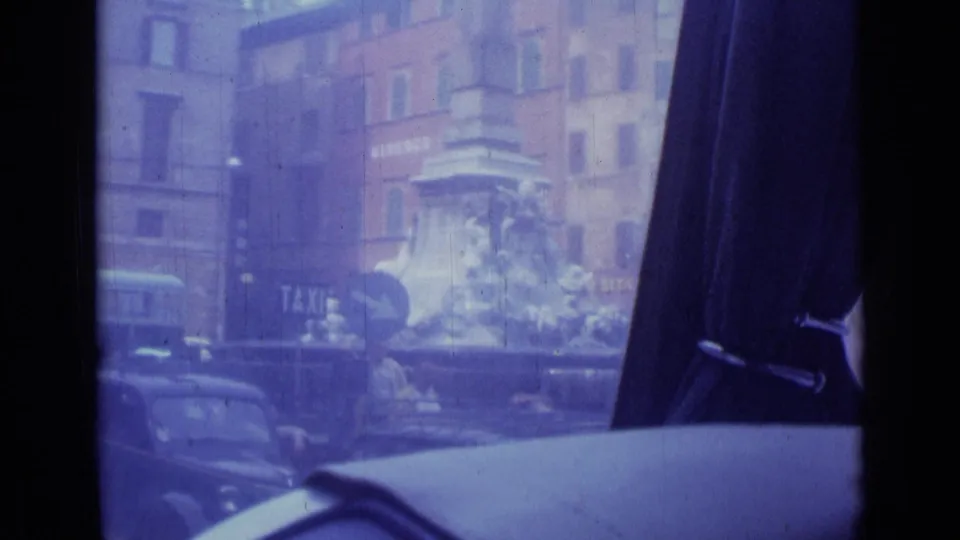 Rome square with a stone monument, visible taxi sign, ALBERGO DEL SOLE building, parked cars, and interior car-window framing.