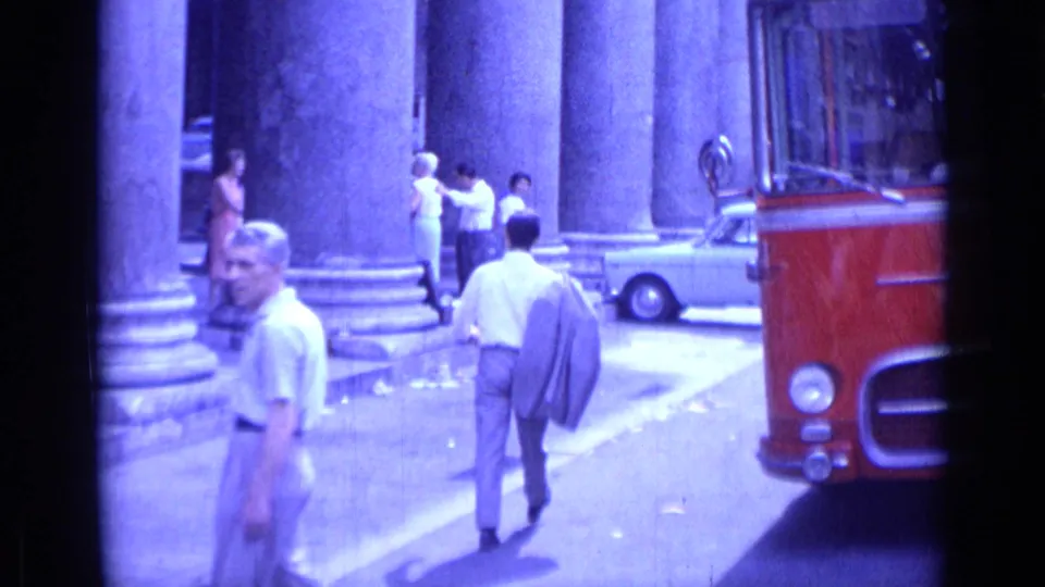 1962 Rome street scene with pedestrians beside massive columns, a red bus at right, and a light-colored car near the curb.