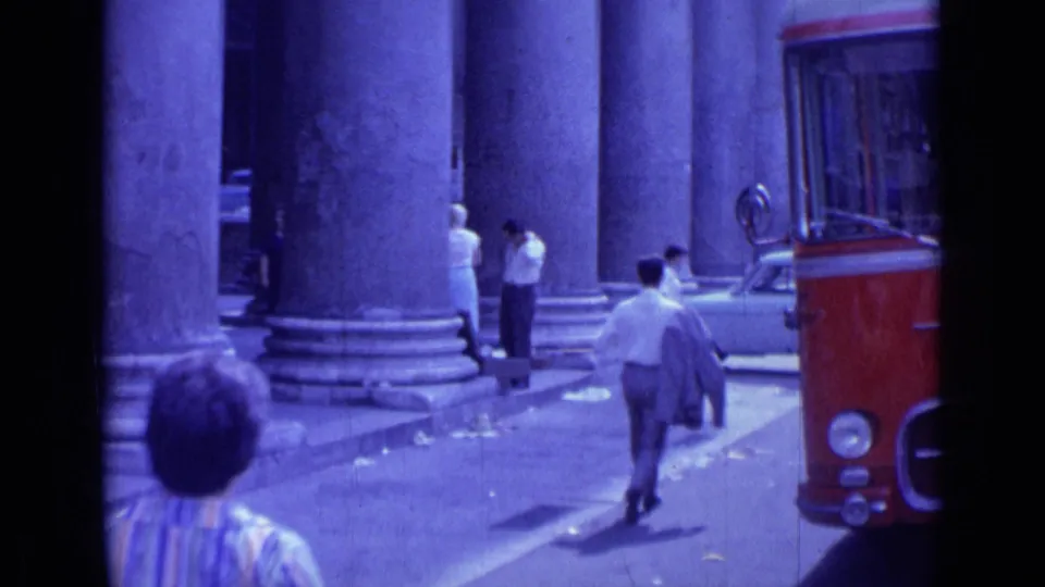 Rome street frame from 1962 showing people walking near monumental columns, with a red bus and pale car at the edge of the road.