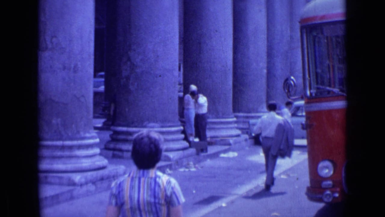 1962 Rome street view with people walking beside massive classical columns and a red trolley car entering the right side of the frame.