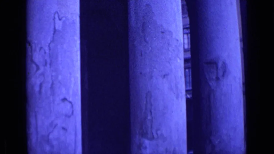 Close handheld 8mm view of weathered classical columns in Rome, with dark portico spaces and limited street detail between the pillars.