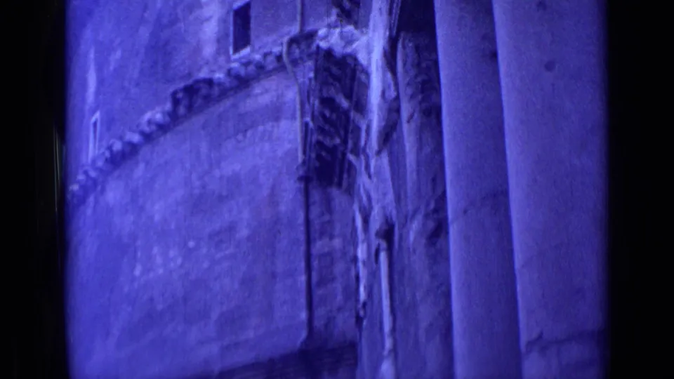 Blue-purple 8mm frame from Rome showing a curved weathered facade section beside massive columns and dark openings.