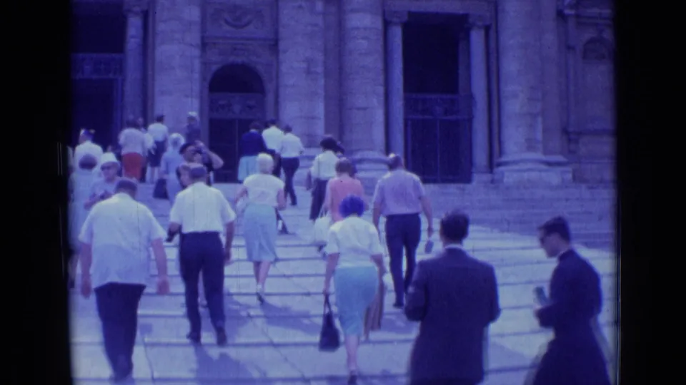 Visitors in mid-century clothing walk across sunlit steps toward a monumental building in Rome, Italy.