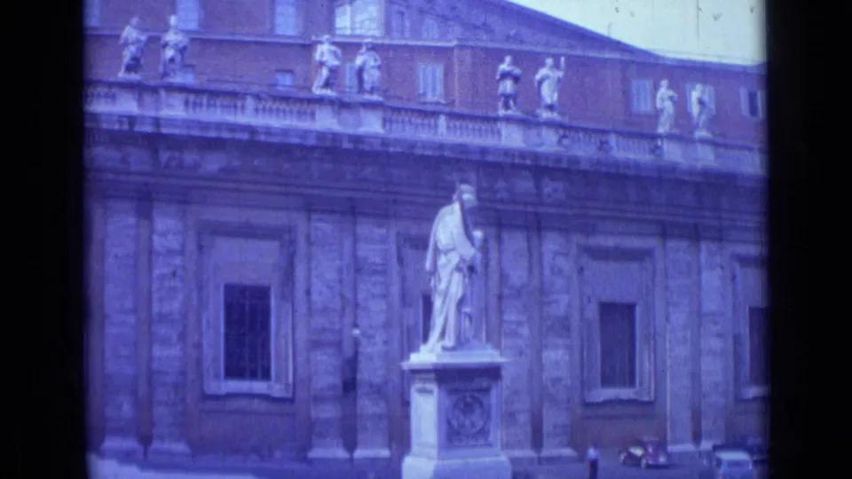A statue on a pedestal appears in front of a grand Roman building facade with roofline figures and windows.
