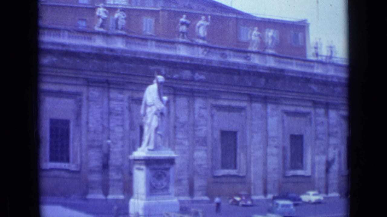 1962 Rome travel-film frame showing a classical facade, rooftop statues, a foreground pedestal statue, and cars along the street.