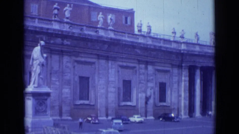 1962 Rome frame from a rightward pan, with a large statue at left, ornate facade details, dark windows, and several cars below.