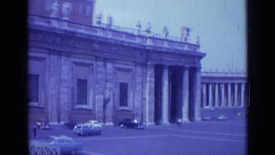 1962 Rome home-movie frame showing a columned entrance, rooftop statues, several cars, and small pedestrians in an open plaza.