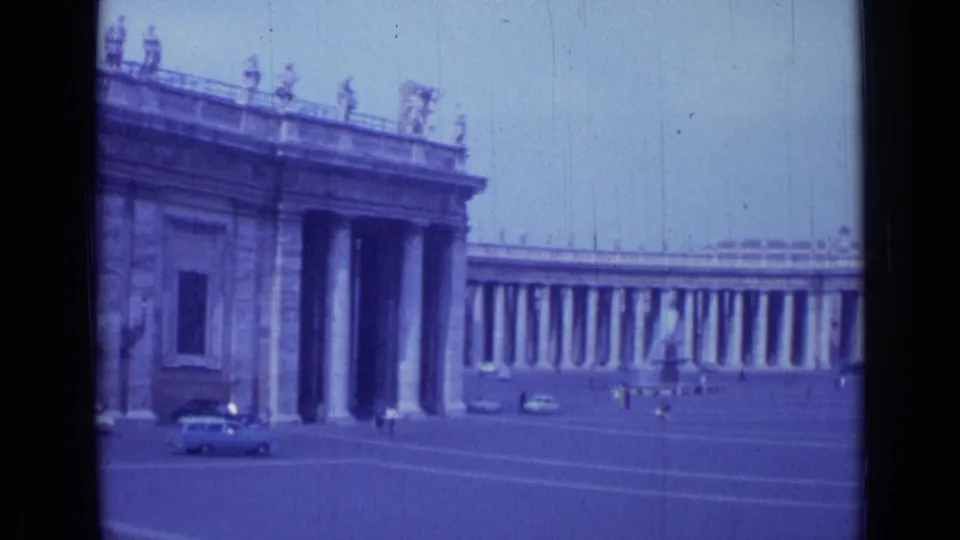 1962 Rome frame showing a columned building entrance, curved colonnade, distant pedestrians, parked cars, and blue-purple film color.