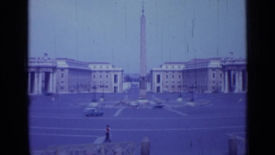 1962 8mm frame of St. Peter's Square with the obelisk, flanking colonnades, passing cars, and a distant pedestrian in red in the foreground.