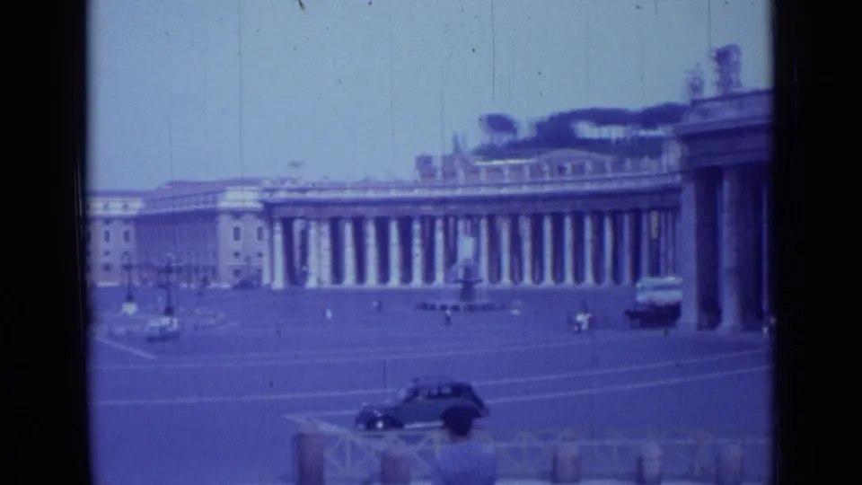 1962 visitor-shot frame in St. Peter's Square showing the colonnades more prominently, with period cars, pedestrians, and foreground barriers.