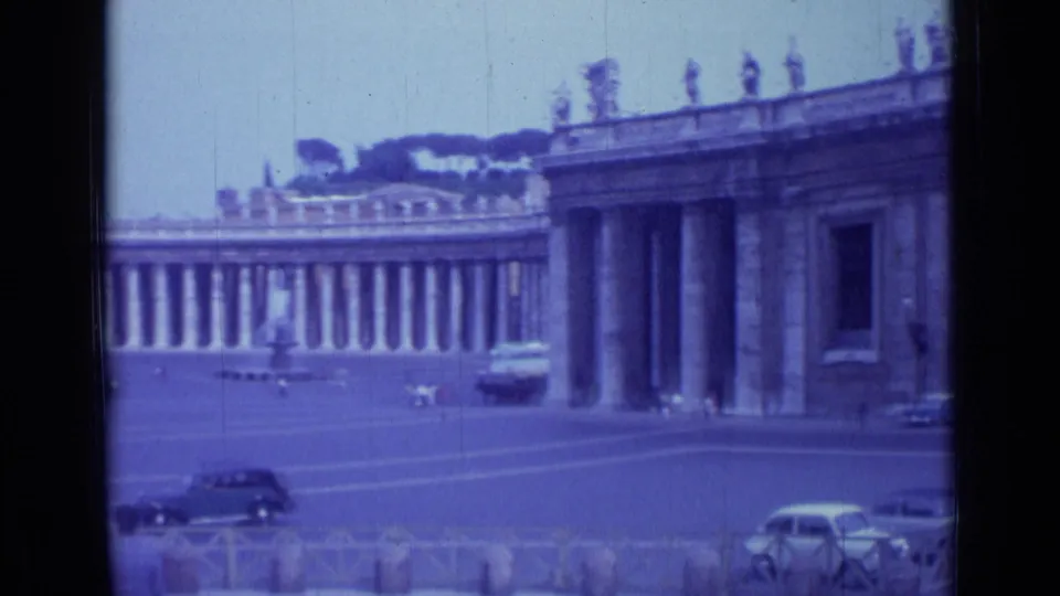 1962 8mm frame of Vatican City's St. Peter's Square, emphasizing the colonnades, foreground cars, low barriers, and small pedestrians.