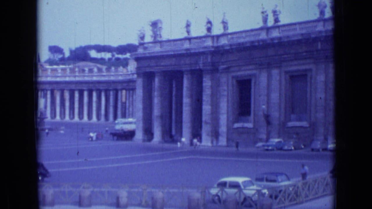 1962 Rome home-movie view of St. Peter's Square, showing the colonnade, rooftop statues, plaza paving, parked cars, and small distant figures.