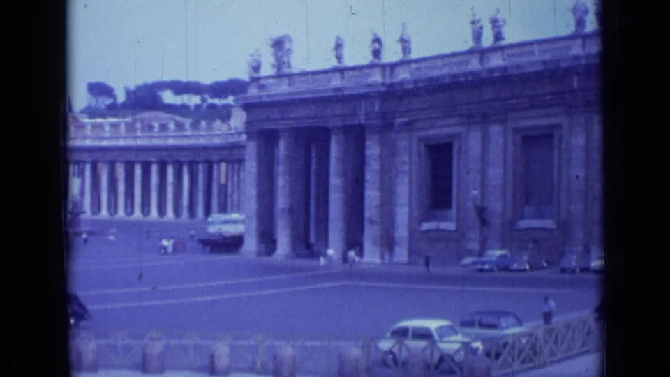 1962 Rome home-movie view of St. Peter's Square, showing the colonnade, rooftop statues, plaza paving, parked cars, and small distant figures.