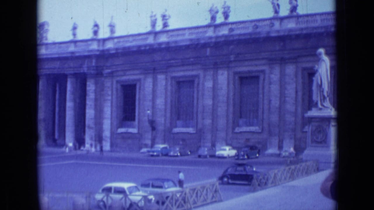 Colonnade facade and statue pedestal beside parked cars and a few distant pedestrians.