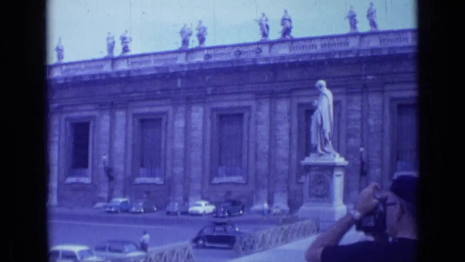 1962 visitor-shot Rome frame showing St. Peter's Square architecture, a pedestal statue, parked cars, railing, and a photographer partly visible in the foreground.