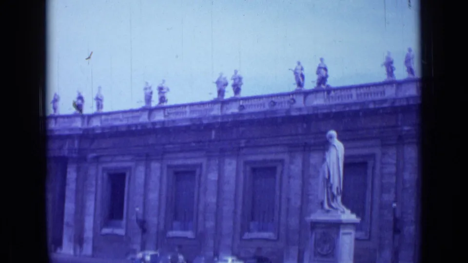 Rome 8mm frame of St. Peter's Square with a pedestal statue, rooftop statues, colonnade wall, parked cars, and soft blue film coloration.