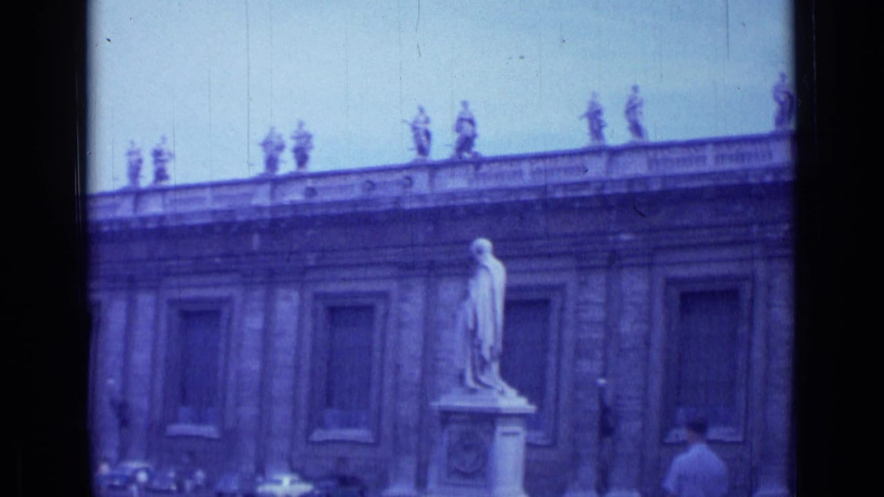 Stone building facade with rooftop statues, a foreground statue, and a tourist crossing the plaza.