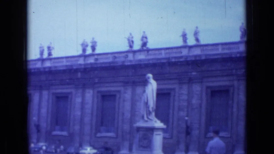 1962 Rome home-movie frame showing a classical stone facade with rooftop statues, a white foreground statue, and a visitor walking below.