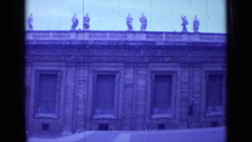 1962 Rome home-movie view of a monumental stone facade with shuttered windows, roofline statues, and low barriers across the plaza.