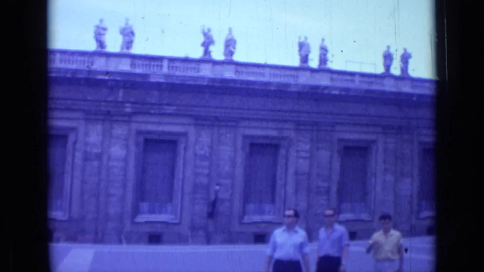 Rome 1962 frame showing several tourists in period clothing walking past a classical building with shuttered windows and rooftop statues.