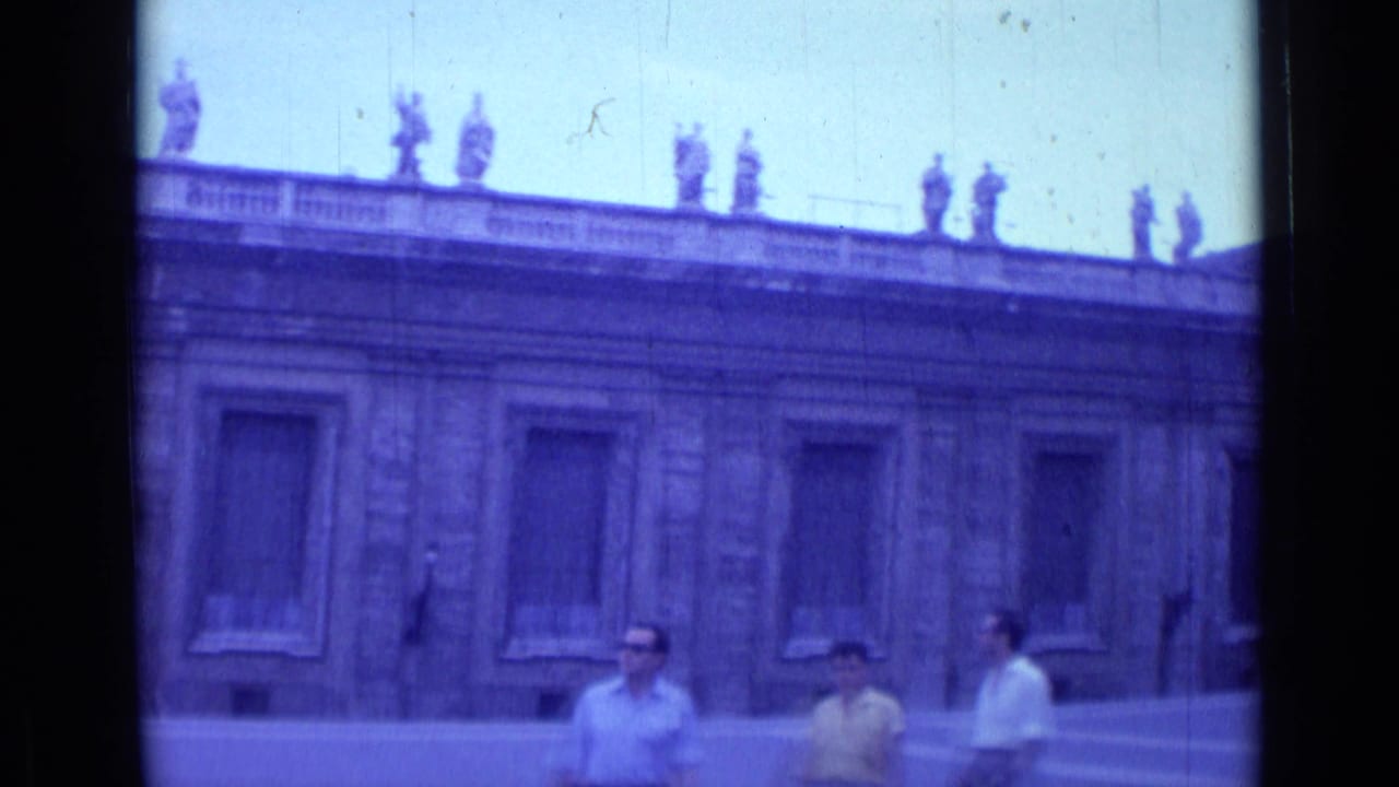 Tourists walk across a paved courtyard in front of a stone church facade with roofline statues.