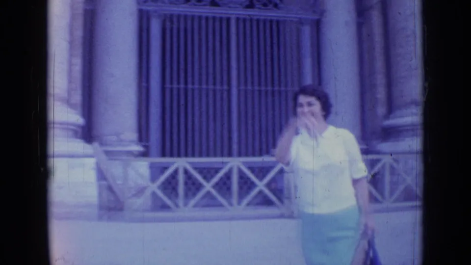 1962 Rome 8mm frame with a woman passing in front of columns, a metal grille, and a low railing.