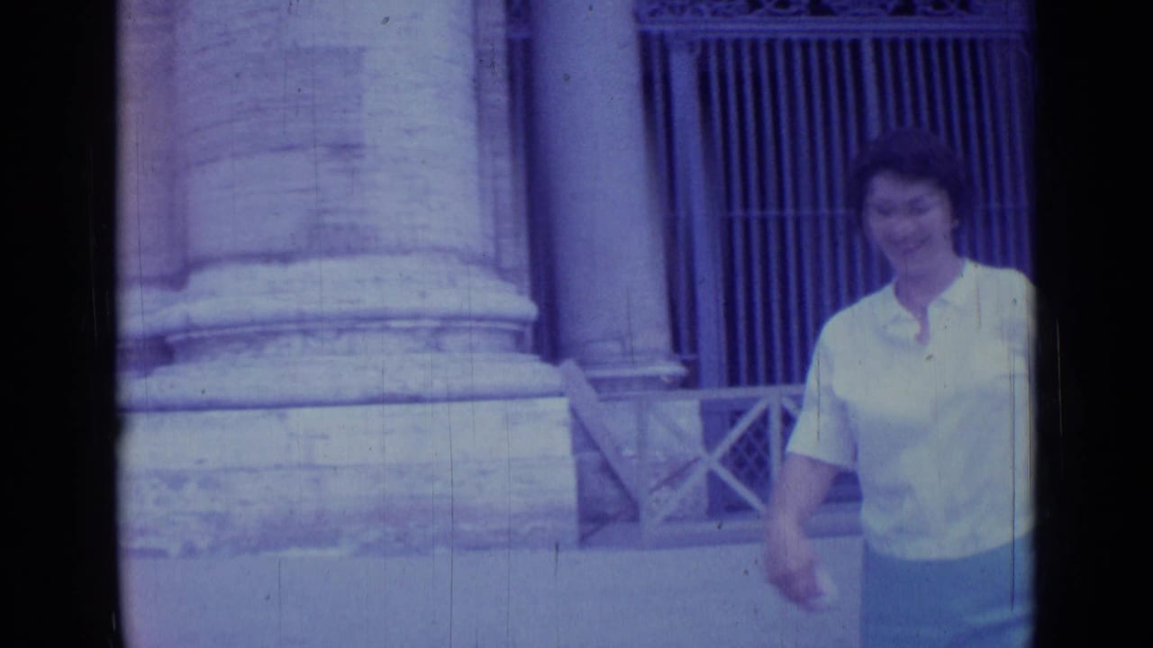 1962 Rome home-movie frame showing a woman in a light blouse and skirt walking past stone columns near a historic entrance.
