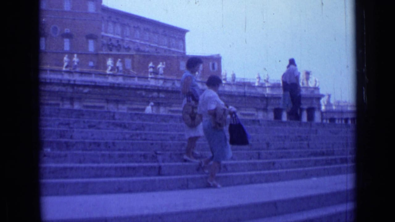 1962 Rome home-movie frame showing tourists in light clothing walking on broad landmark steps beneath masonry buildings and statues.