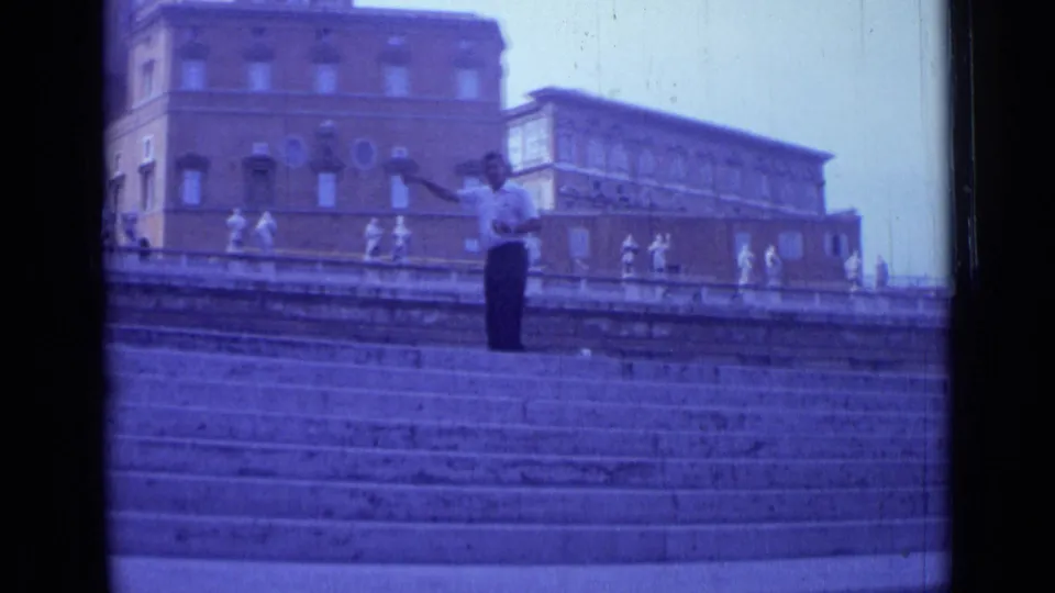 1962 Rome home-movie frame showing a man gesturing on broad stone steps below a terrace with statues and masonry buildings.