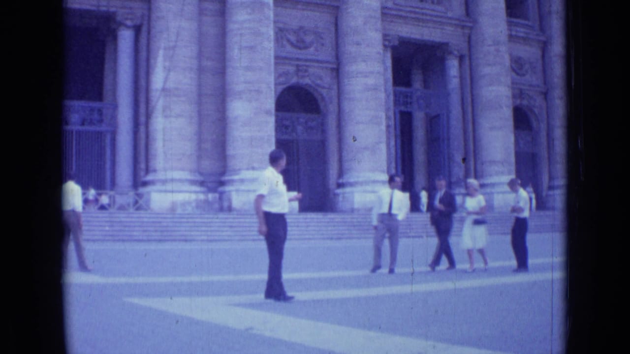 In 1962 Rome footage, a man in a white shirt is seen near broad steps and massive columns as pedestrians move nearby.