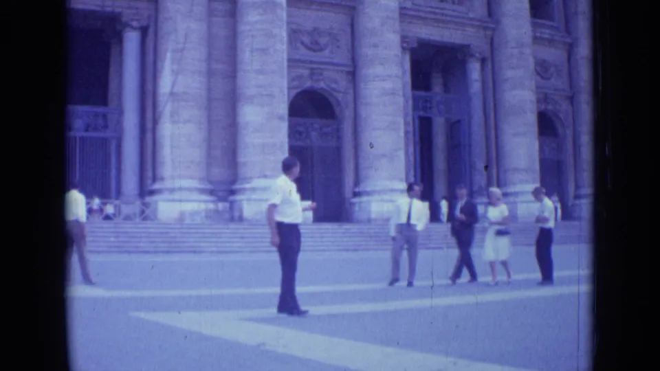 In 1962 Rome footage, a man in a white shirt is seen near broad steps and massive columns as pedestrians move nearby.