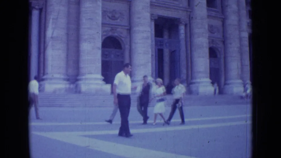 The Rome street scene shows several adults crossing before the columned stone building as the man in white moves nearer to camera.