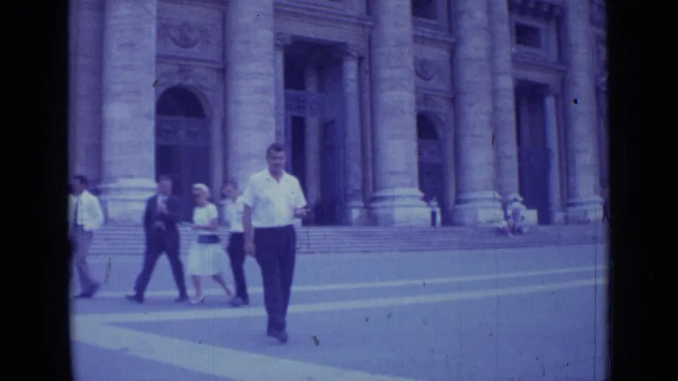 A man in a short-sleeved white shirt approaches the camera in a 1962 Rome plaza, with pedestrians and stone steps behind him.