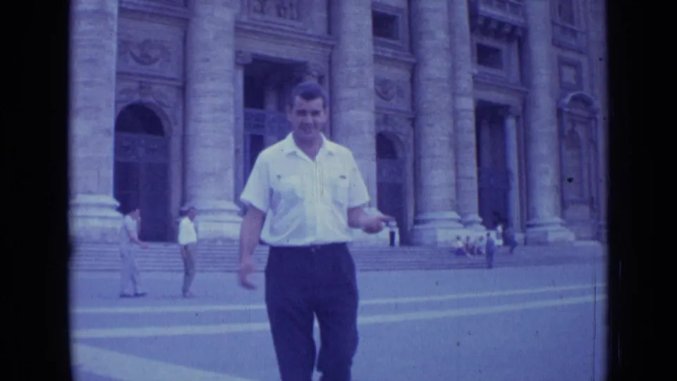 The man continues walking toward the camera in a 1962 Rome plaza, with pedestrians and ornate stonework in the background.