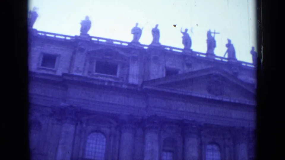Rome 1962 home-movie frame of St. John Lateran's upper facade, showing statues, a triangular pediment, arched windows, and aged 8mm color shift.