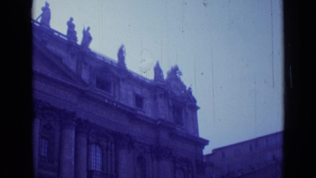 Stone building facade with roofline statues and a bright sky, seen in blue-tinted 8mm footage.