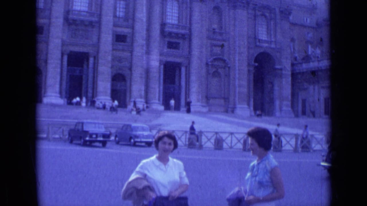 Two women stand in a large plaza before a columned cathedral facade with parked cars nearby.