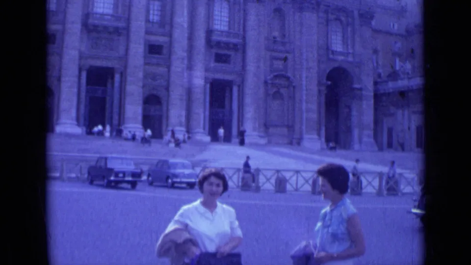 Two women tourists converse in a Rome plaza in 1962, with a large columned facade, parked cars, and pedestrians behind them.