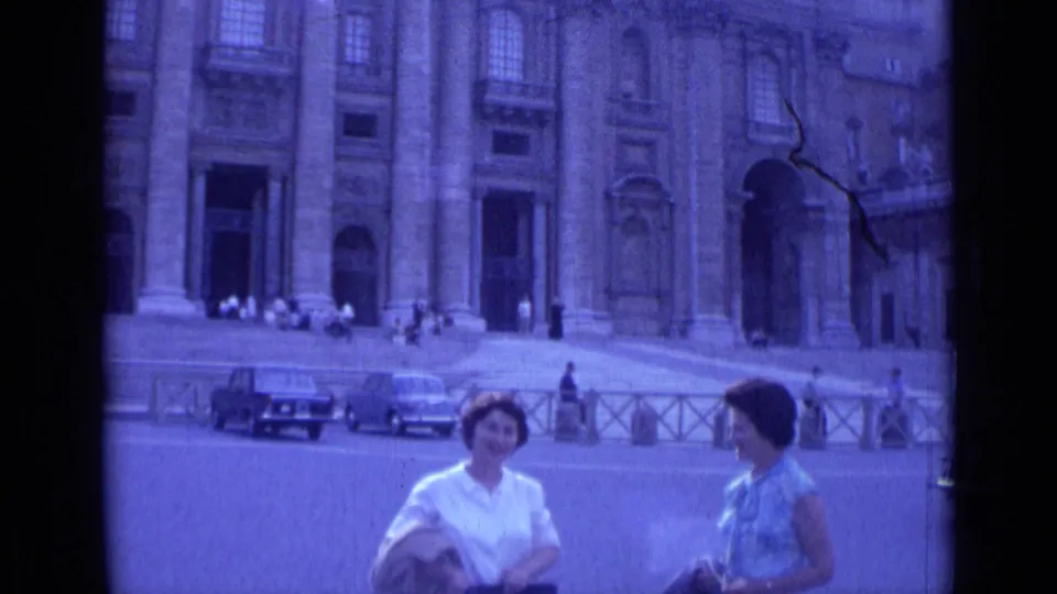 Rome 1962 frame of two women tourists in a plaza before a monumental cathedral-like facade, with cars and railings in the background.