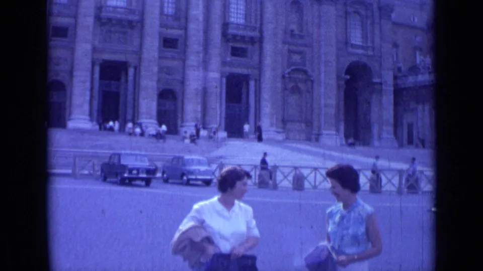 Two women tourists face each other in a Rome plaza in 1962, framed against a large stone facade with cars and pedestrians nearby.