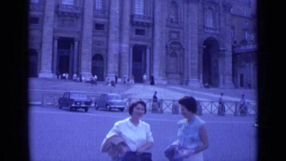 Two women stand chatting in Rome in 1962, with St. Peter's Basilica, parked cars, railings, and pedestrians visible behind them.